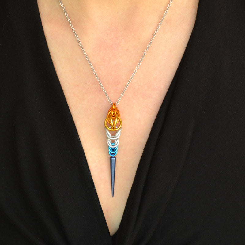 Close-up of a chainmaille necklace on a woman’s neck. The necklace is long and narrow, made in the colors of the AroAce pride flag. The chainmaille weave is orange at top, transitioning to white then blue, with a dark blue acrylic spike at the bottom. The woman is wearing a black V-neck blouse.