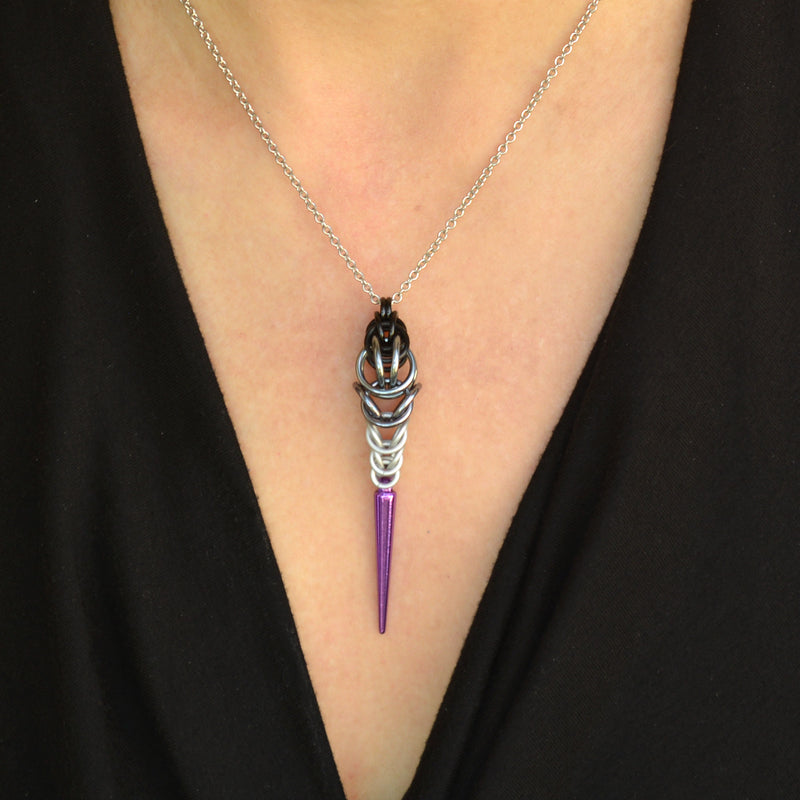Close-up of a chainmaille necklace on a woman’s neck. The necklace is long and narrow, made in the colors of the Asexual pride flag. The chainmaille weave is black at top, transitioning to grey and then white, with a light purple acrylic spike at the bottom. The woman is wearing a black V-neck blouse.