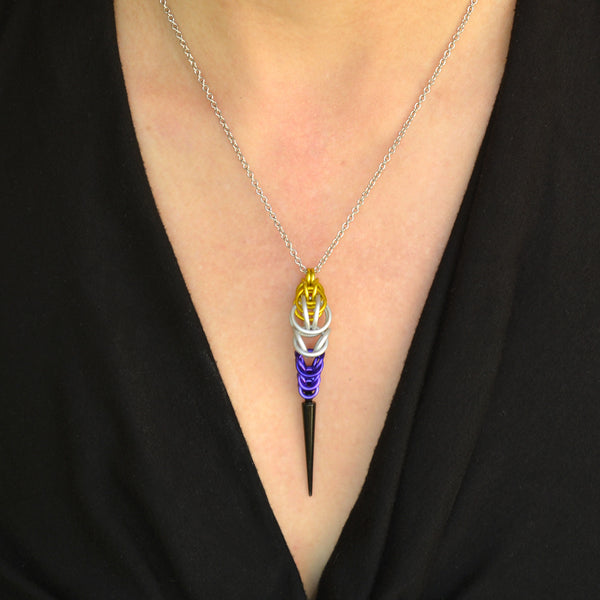 Close-up of a chainmaille pendant on a thin silver chain on a woman’s neck. The pendant is long and narrow, made in the colors of the nonbinary pride flag. The chainmaille weave is yellow at top, transitioning to white and then purple, with a black acrylic spike at the bottom. The woman is wearing a black V-neck top.