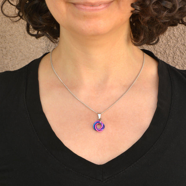 Chainmaille artisan Rebeca Mojica wearing a black v-neck shirt modeling her bi pride small swirl necklace on a stainless steel chain. The necklace has 5 intertwined links in pink, blue and lilac to match the bisexual pride flag colors. The background is a textured beige wall.