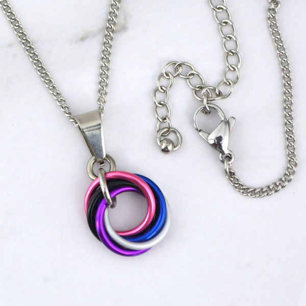 Close-up of a small genderfluid pride chainmaille pendant resting on a marble surface. The pendant is a vortex of 5 intertwined rings in the colors of the genderfluid pride flag: pink, white, violet, blue and black. The links are joined by a steel ring and attached to a smooth steel bail. A steel chain runs through the bail, with the lobster clasp shown on the right.