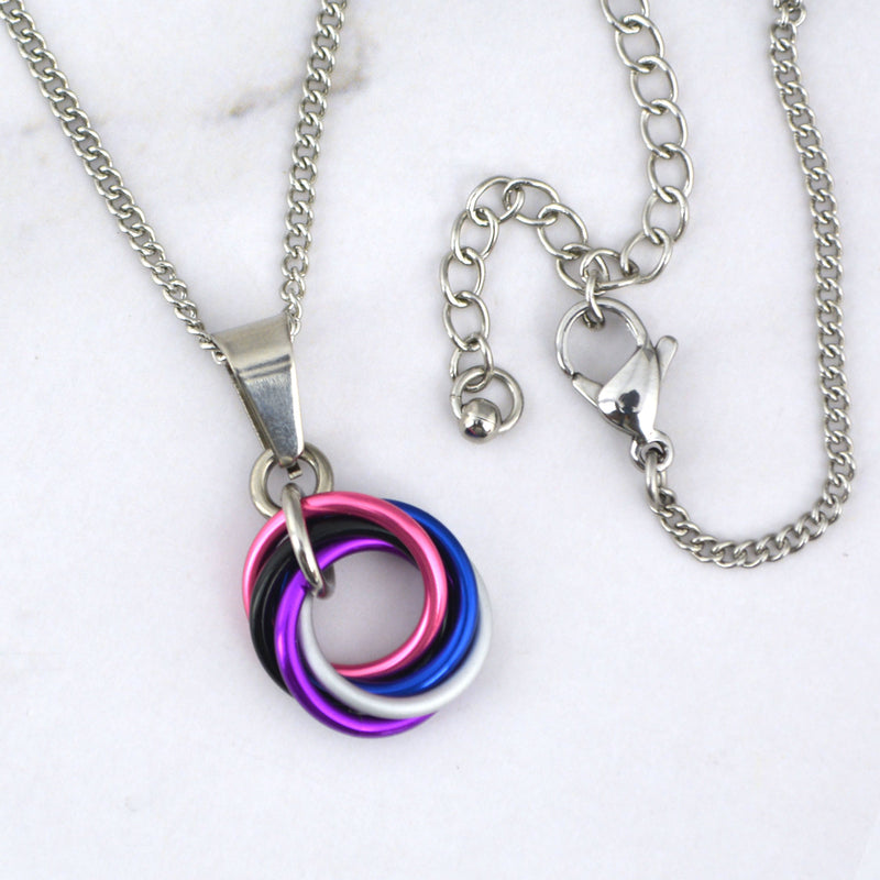 Close-up of a small genderfluid pride chainmaille pendant resting on a marble surface. The pendant is a vortex of 5 intertwined rings in the colors of the genderfluid pride flag: pink, white, violet, blue and black. The links are joined by a steel ring and attached to a smooth steel bail. A steel chain runs through the bail, with the lobster clasp shown on the right.