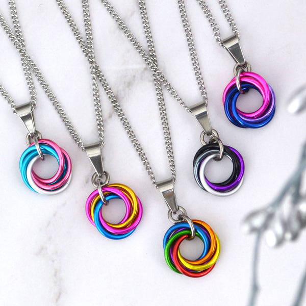 Five necklaces with different colored woven ring pendants. The pendants are in the colors of the transgender, pansexual, rainbow, asexual, and bisexual pride flags. The necklaces are on a white marble surface.
