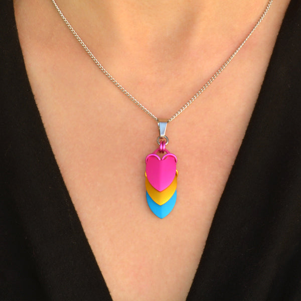 Closeup of chevron-style pansexual pride pendant on silver chain. Pendant is a series of stacked leaf-shaped metal components in anodized aluminum, each in a color of the pan flag: from top to bottom pink, gold and blue. Pendant shown worn on a light-skinned person wearing a black top.
