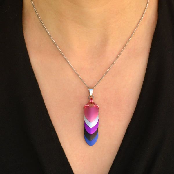 Closeup of chevron-style gender-fluid pride pendant on silver chain. Pendant is a series of stacked leaf-shaped metal components in anodized aluminum, each in a color of the genderfluid flag: from top to bottom pink, white, purple, black and blue. Pendant shown worn on a light-skinned person wearing a black top.
