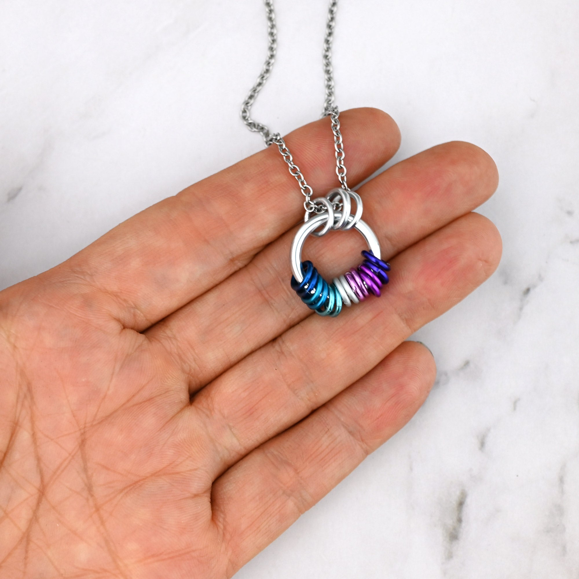 The Berrisexual pride coil necklace held on fingers for scale. The multiple colored rings are clustered at the bottom in a way that shows they move freely and independently.
