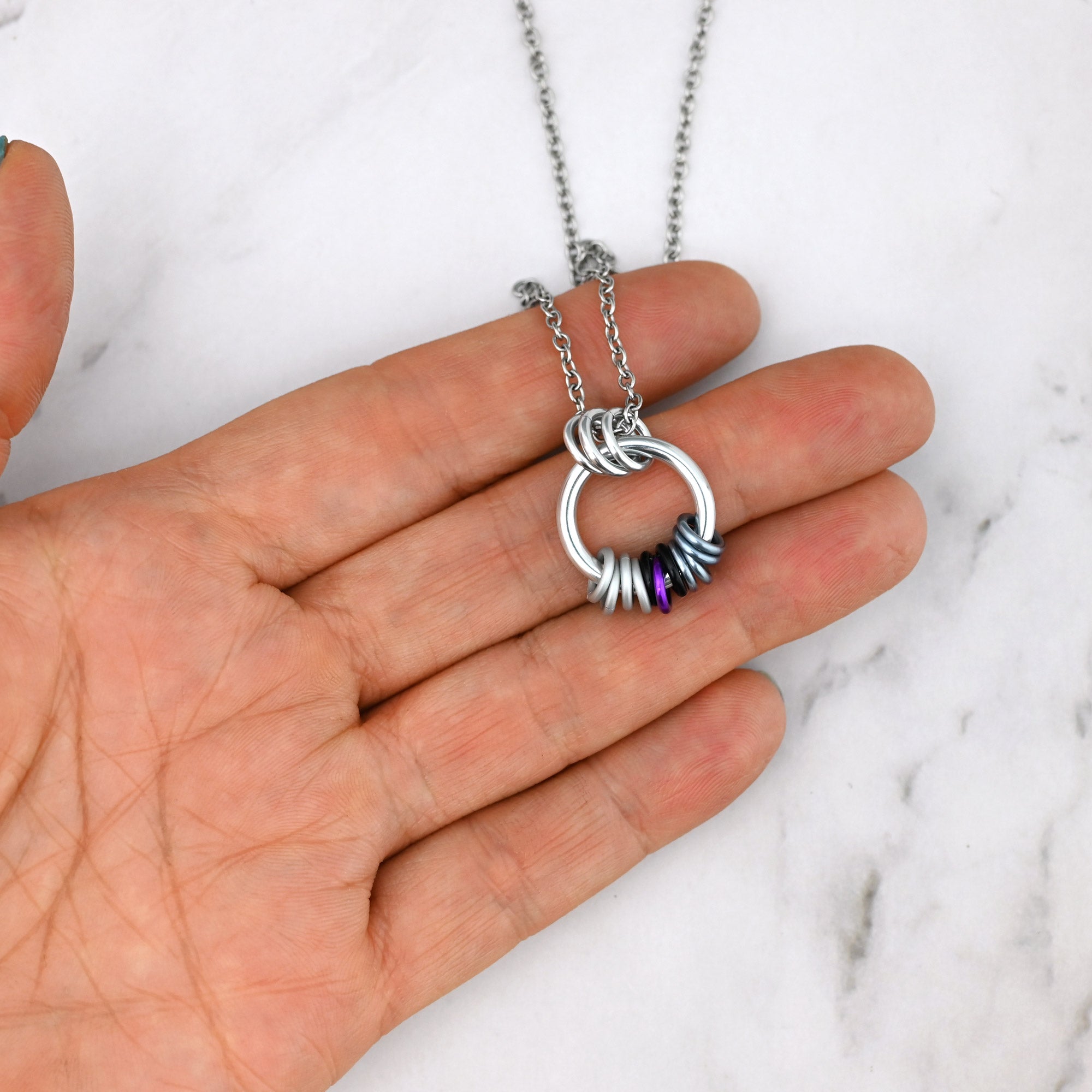  The Demisexual coil necklace held against a person's fingers, illustrating the small, discreet nature of the 0.75-inch pendant and the tactile arrangement of the moveable rings.