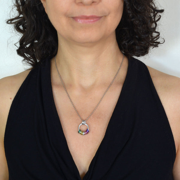 Rebeca Mojica, a chainmaille artisan, modeling the Genderfae Pride Coil Fidget Necklace while wearing a black top. The image subtly emphasizes the necklace's small, discreet size, featuring green, light green, light yellow, white, lilac, and lavender rings that best match the Genderfae Pride flag.