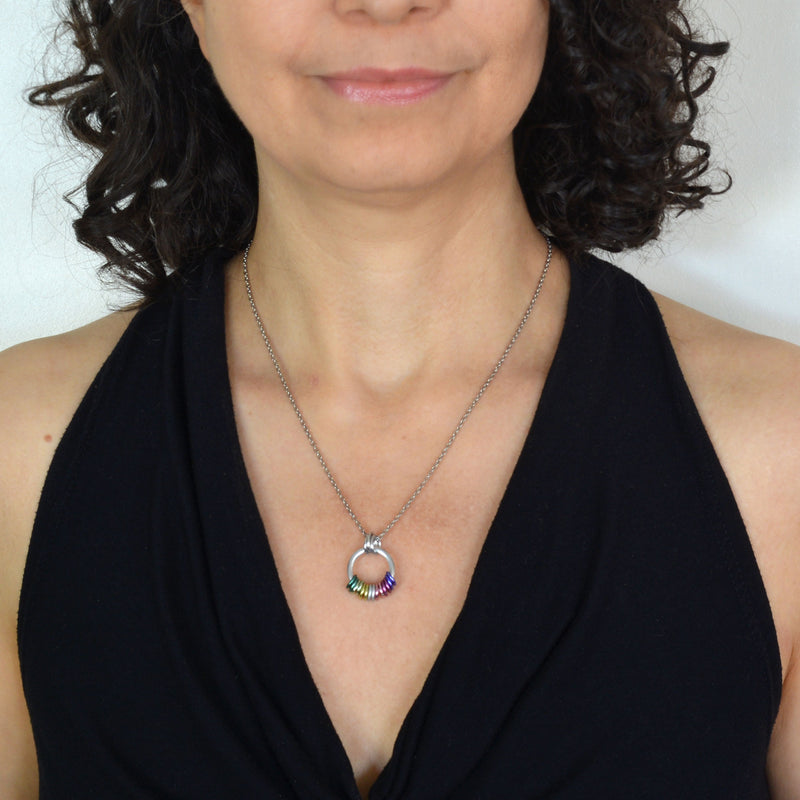 Rebeca Mojica, a chainmaille artisan, modeling the Genderfae Pride Coil Fidget Necklace while wearing a black top. The image subtly emphasizes the necklace's small, discreet size, featuring green, light green, light yellow, white, lilac, and lavender rings that best match the Genderfae Pride flag.