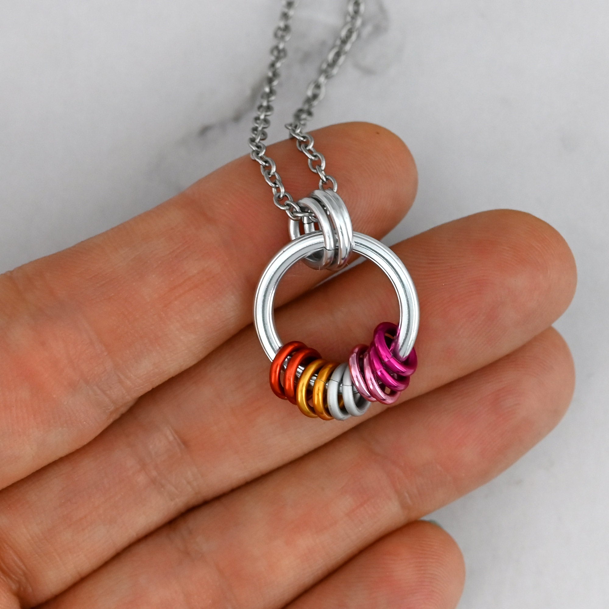 A hand holds the minimalist Lesbian Pride coil necklace, demonstrating its small size (about 0.75 inches wide). The sunset-colored rings sit together at the base of the hoop, and their loose alignment conveys that they slide and rotate freely for fidgeting.