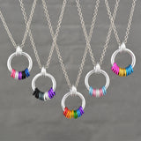A group of five distinct Pride Coil Fidget Necklaces, each showcasing different flag colors (from left: Genderfluid, Asexual, Rainbow, Transgender, Pansexual). The silver coil pendants with their freely spinning aluminum rings are displayed against a neutral grey background.
