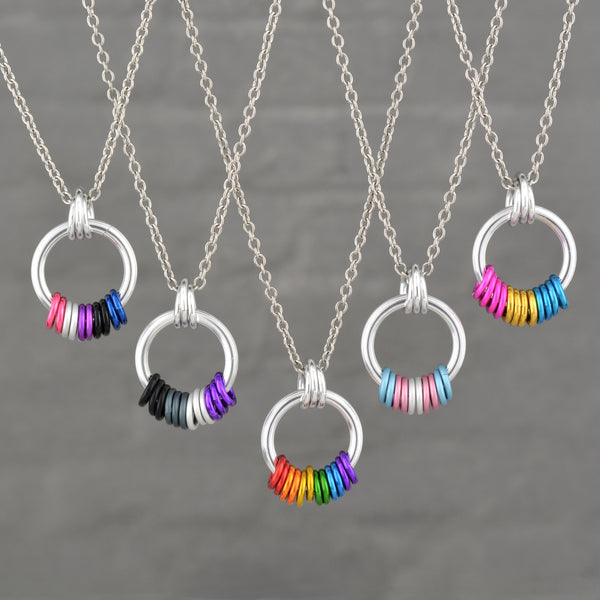 A group of five distinct Pride Coil Fidget Necklaces, each showcasing different flag colors (from left: Genderfluid, Asexual, Rainbow, Transgender, Pansexual). The silver coil pendants with their freely spinning aluminum rings are displayed against a neutral grey background.
