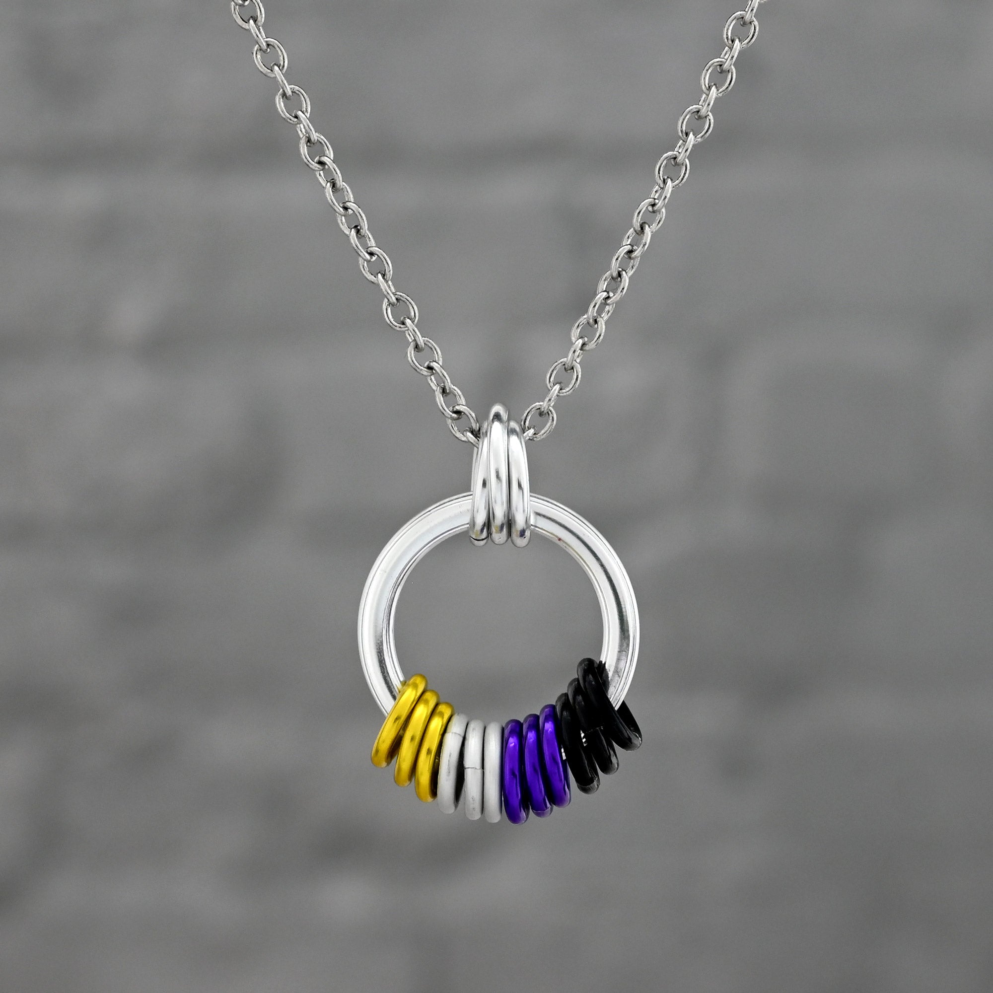 A silver circular pendant featuring a cluster of rings in the Nonbinary Pride flag colors: yellow, white, purple, and black. The pendant is suspended by a stainless steel chain.