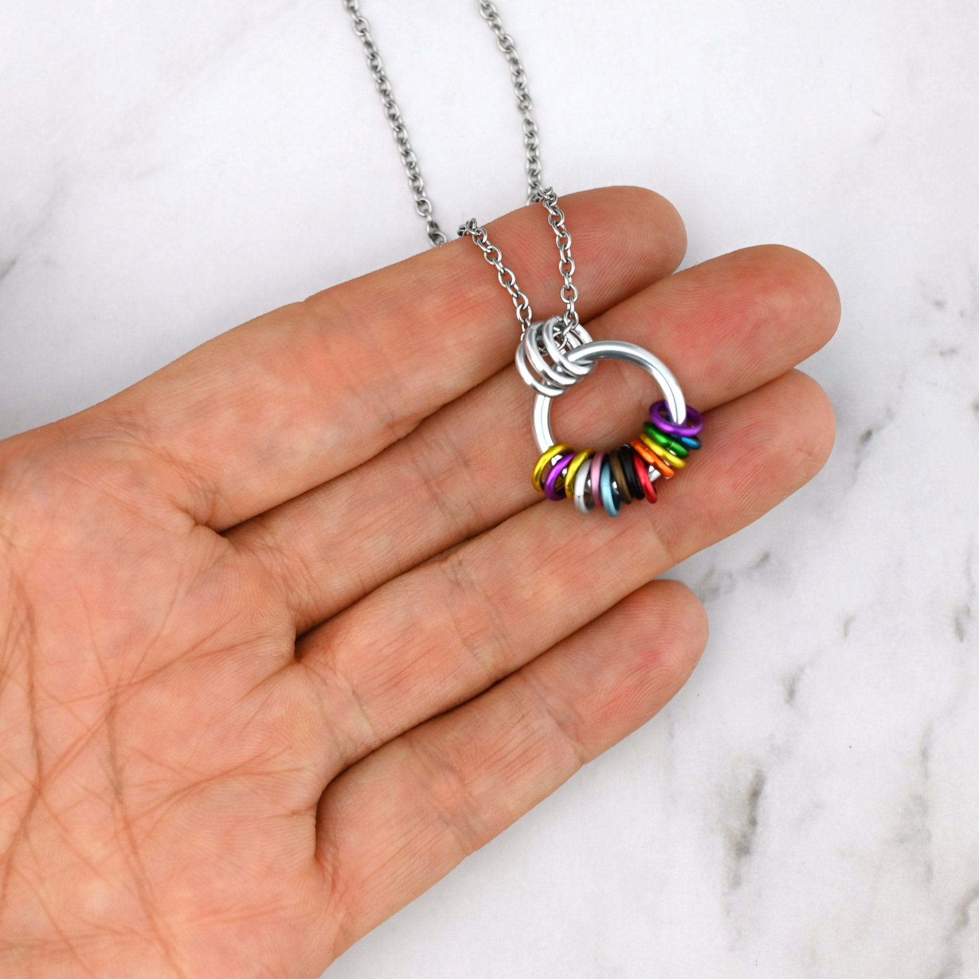  The Intersex-Inclusive Progress pride coil necklace held on fingers for scale. The multiple colored rings are clustered at the bottom in a way that shows they move freely and independently.