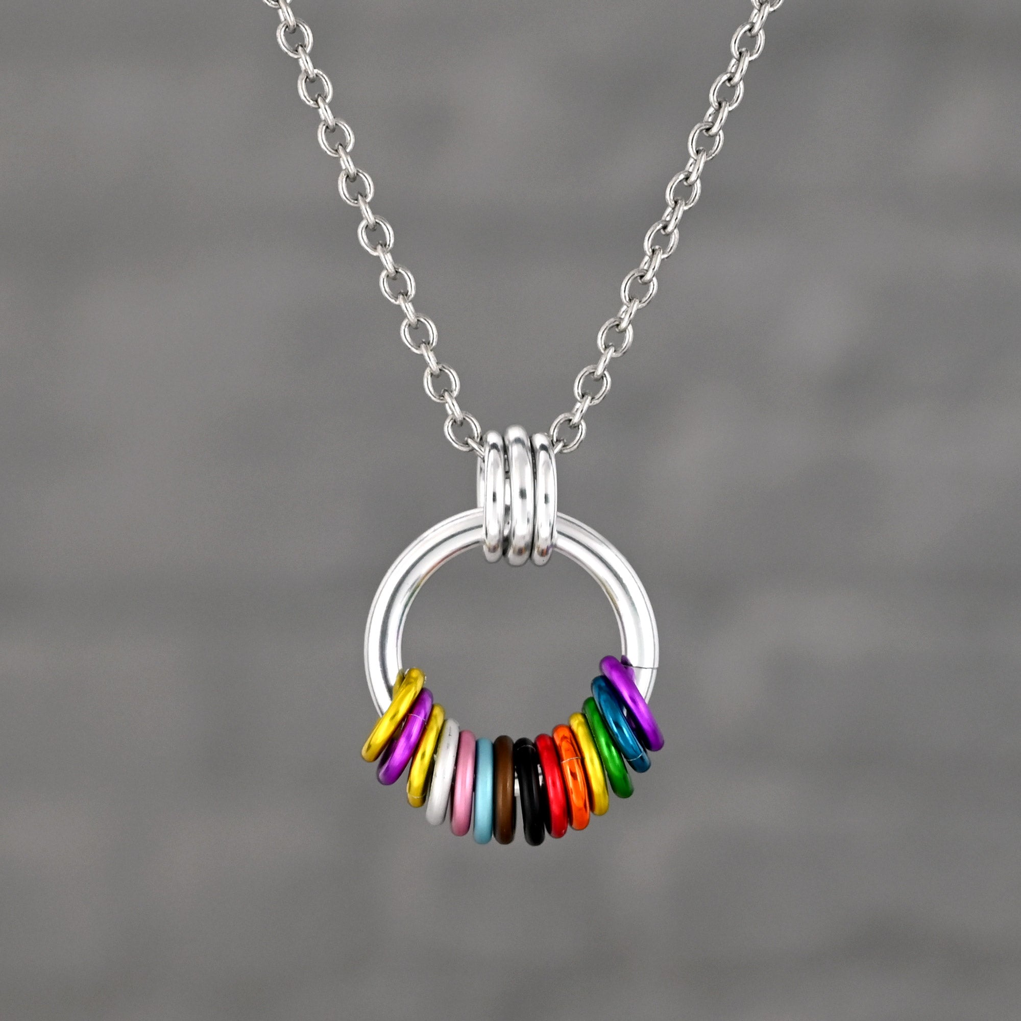 A silver circular pendant featuring a spectrum of colored rings representing the intersex-inclusive progress flag, from left to right: yellow, purple, yellow, purple, white, pink, blue, brown, black, and the traditional lgbtq rainbow colors.