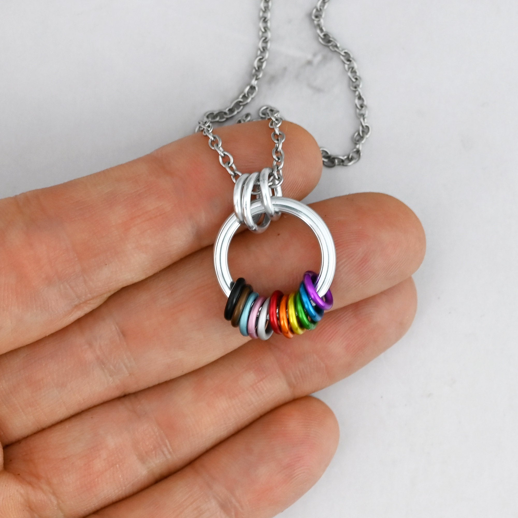 The Progress Pride coil necklace held against a person's fingertips, highlighting its compact, lightweight design and the tactile, free-spinning feature of the multiple colored rings.