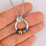 A close-up view of the Progress Pride Coil Fidget Necklace gently held between fingertips. The image highlights the silver ring and the freely spinning black, brown, blue, pink, and rainbow-colored aluminum rings of the Progress flag.