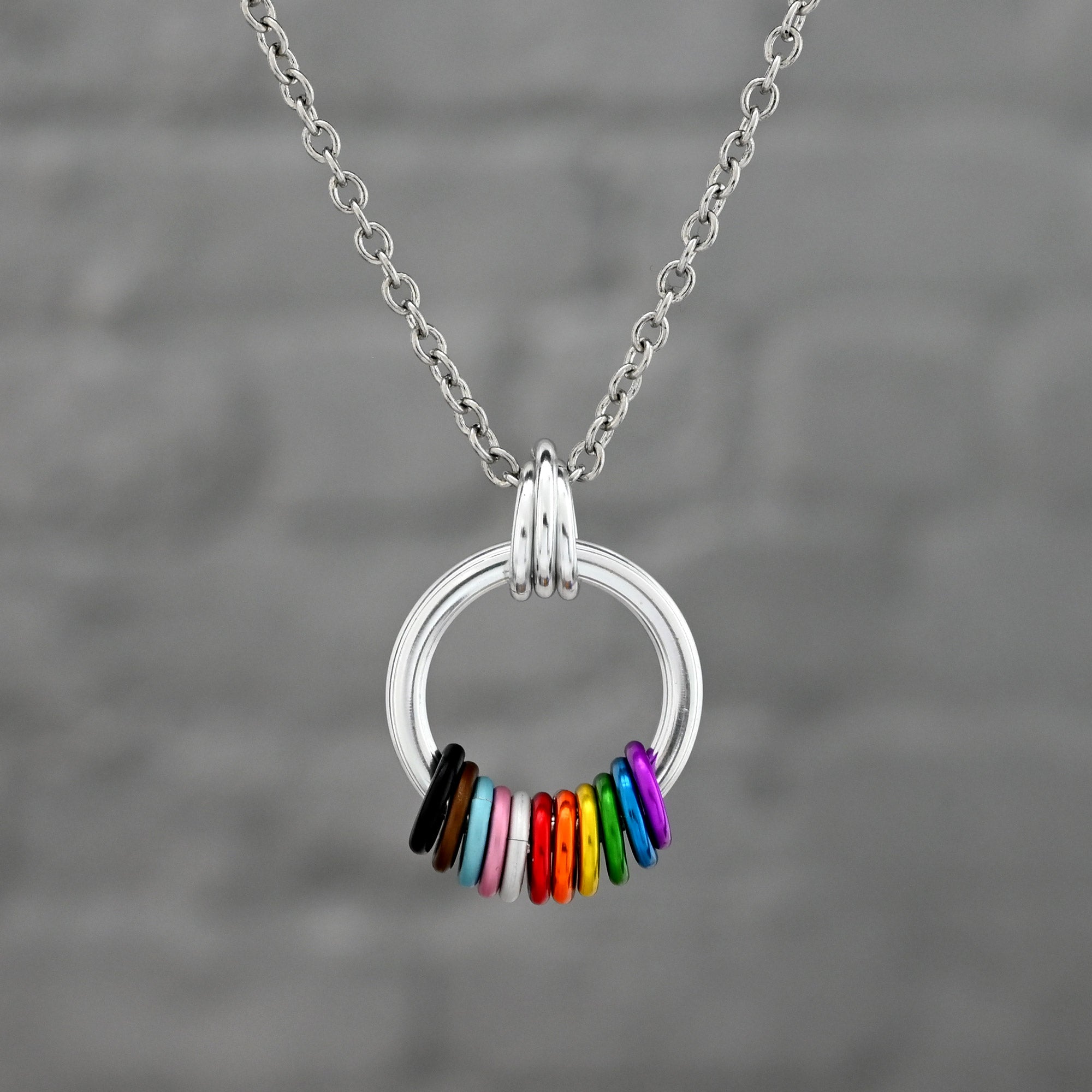 A silver circular pendant on a chain featuring a spectrum of colored rings representing the Progress Pride flag. The rings include black, brown, light blue, pink, and white, followed by the traditional rainbow colors.