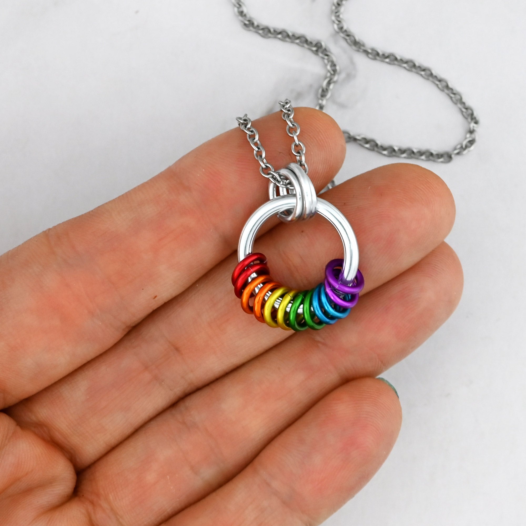 A hand holds the rainbow coil pendant to show its small scale, measuring approximately 0.75 inches wide. The colorful rings sit loosely at the base of the silver hoop, highlighting the tactile, fidget-friendly design.