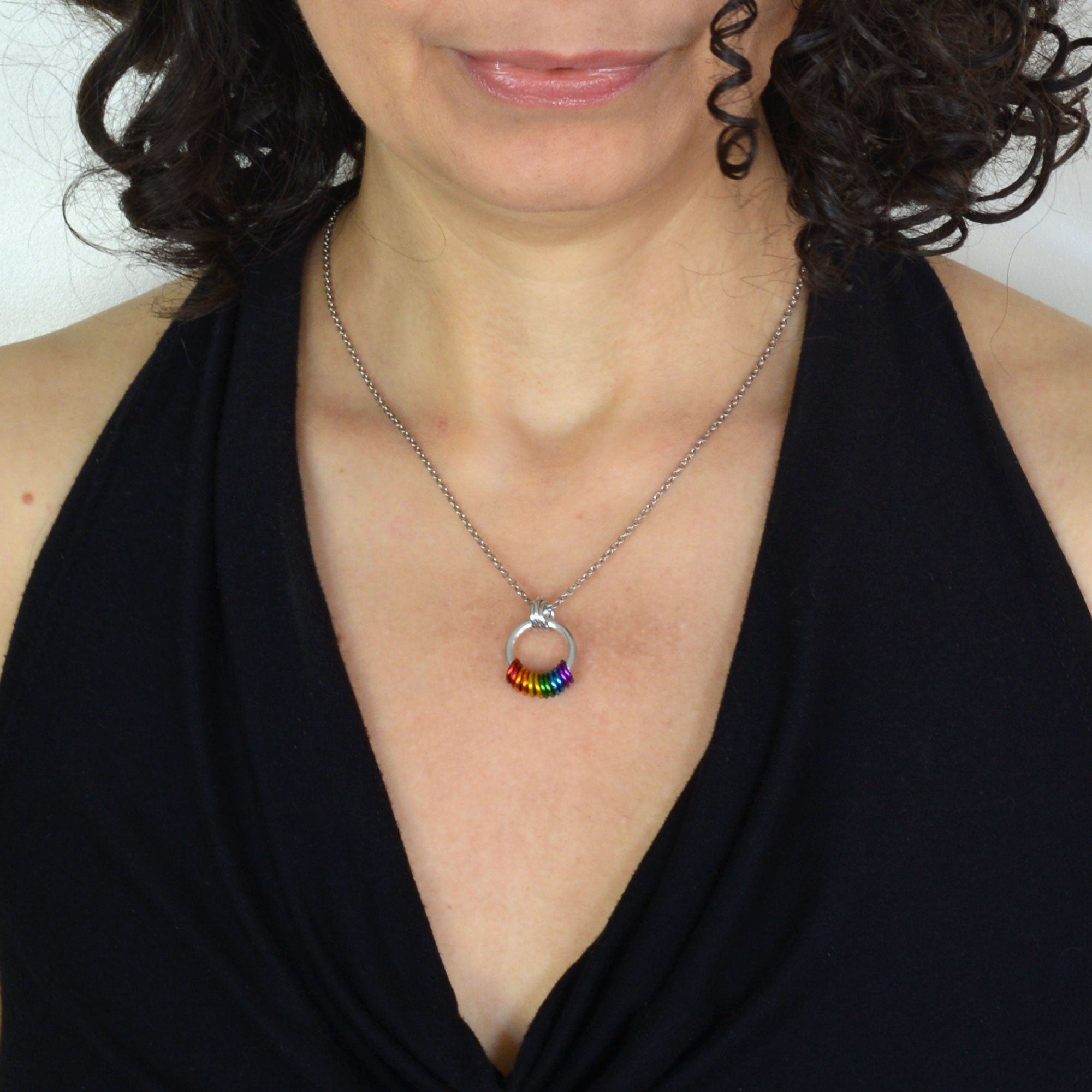 Woman with curly brown hair wearing the rainbow pride small coil necklace and a black halter top. The chainmaille necklace is about the size of a quarter, featuring 12 links in the color of the lgbtq rainbow pride flag hanging from a main silver color link.