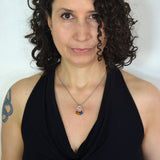 Rebeca Mojica, a chainmaille artisan, modeling the Rainbow Pride Coil Fidget Necklace. She wears a black top and has a subtle tattoo on her arm. Her shoulder-length curly dark brown hair frames her face as she looks directly at the camera with a slight smile. The necklace features bright rainbow-colored rings, worn discreetly.

