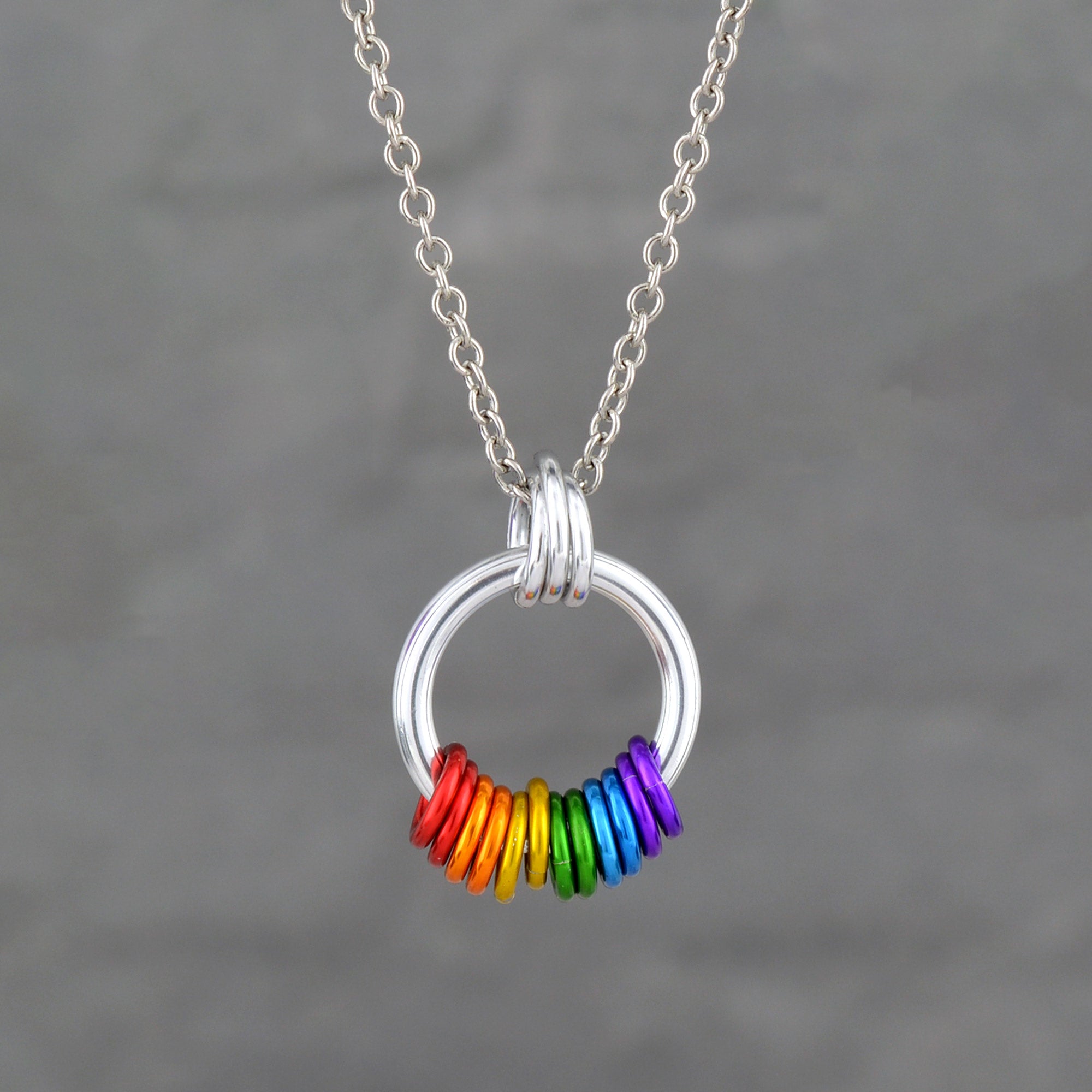 A modern, minimalist silver-tone circular pendant hanging from a stainless steel chain. The bottom of the circle features a series of small, free-spinning aluminum rings in a vibrant rainbow sequence: red, orange, yellow, green, blue, and purple.