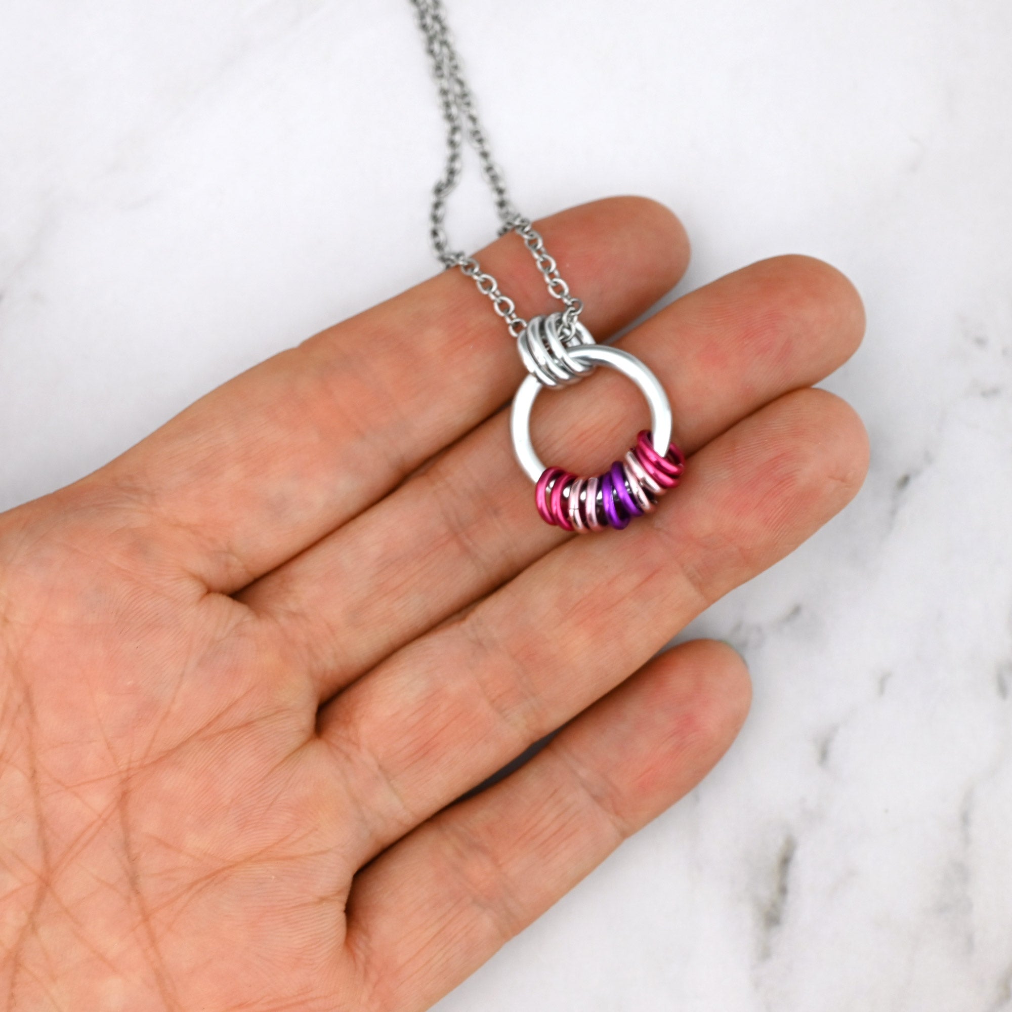 A close-up scale shot of the Sapphic pride chainmaille pendant held in a hand. The pink and purple rings are stacked along the bottom of the silver circle, sitting in a coil-like arrangement, but spaced out slightly to indicate they are free to move and slide along the main silver link.