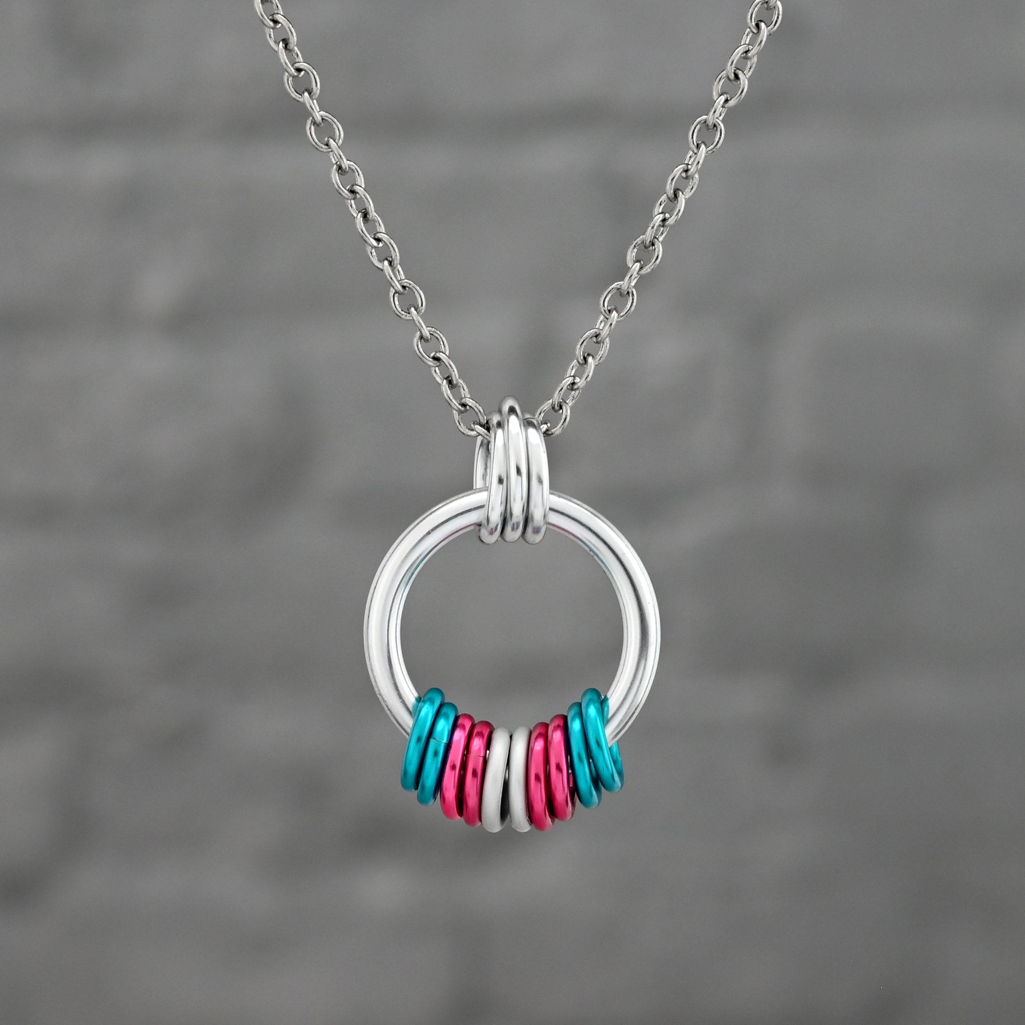 A minimalist silver hoop pendant featuring aluminum rings in the Transgender Pride flag colors: light blue, pink, and white. The rings are arranged symmetrically on the bottom of the hoop.