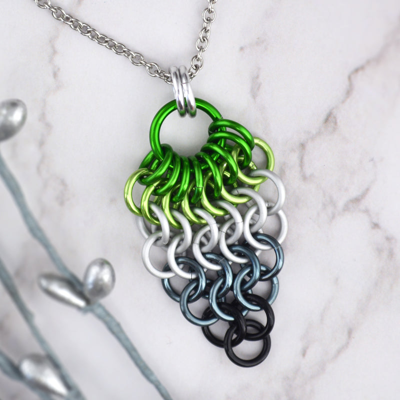 Closeup of short aromantic pride chainmaille mesh pendant by Rebeca Mojica displayed on a marbled surface. Pendant is reverse teardrop shape with a large green ring at top. From that ring, a row of 8 green rings hang and are connected to a row of 7 light green rings beneath them. Each row gets smaller and smaller: 6 white rings, 5 white rings, 4 grey rings, 3 grey rings, 2 black rings and 1 black ring