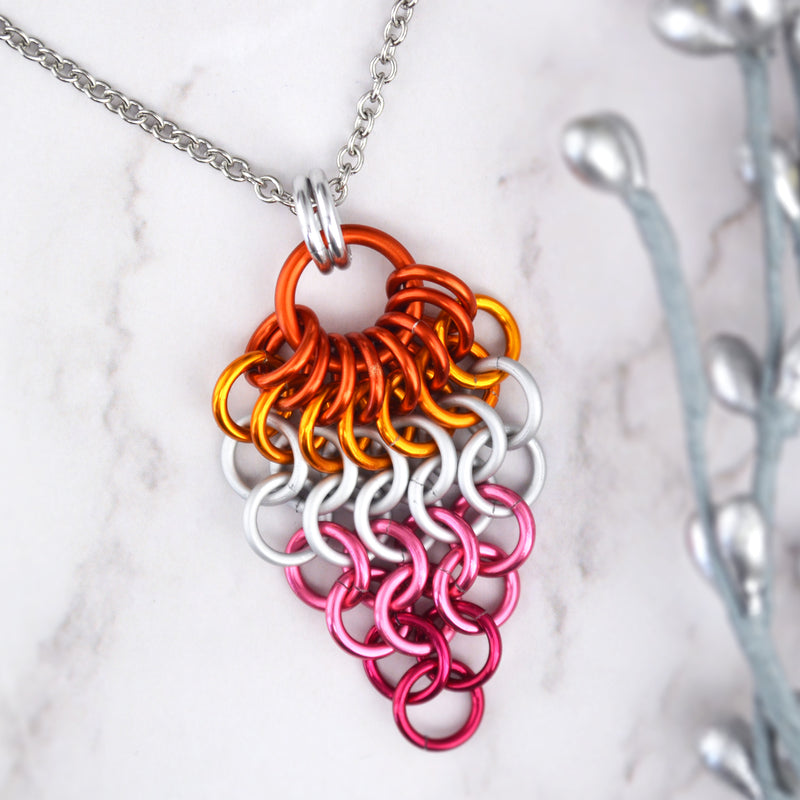 Closeup of short lesbian pride chainmaille mesh pendant by Rebeca Mojica displayed on a marbled surface. Pendant is reverse teardrop shape with a large dark orange ring at top. From that ring, a row of 8 dark orange rings hang and are connected to a row of 7 bright orange rings beneath them. Each row gets smaller and smaller: 6 white rings, 5 white rings, 4 pink rings, 3 pink rings, 2 fuchsia rings and 1 fuchsia ring. 