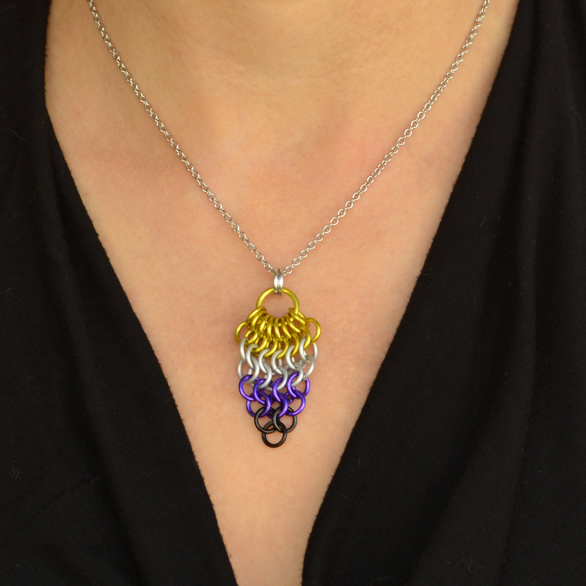 Closeup of small nonbinary pride chainmaille mesh pendant by Rebeca Mojica being worn on a light skinned person with a plunging V-neck black top. Pendant is reverse teardrop shape with a large bright yellow ring at top. From that ring, a row of 8 yellow rings hang and are connected to a row of 7 yellow white rings beneath them. Each row gets smaller and smaller: 6 white rings, 5 white rings, 4 purple rings, 3 purple rings, 2 black rings and 1 black ring
