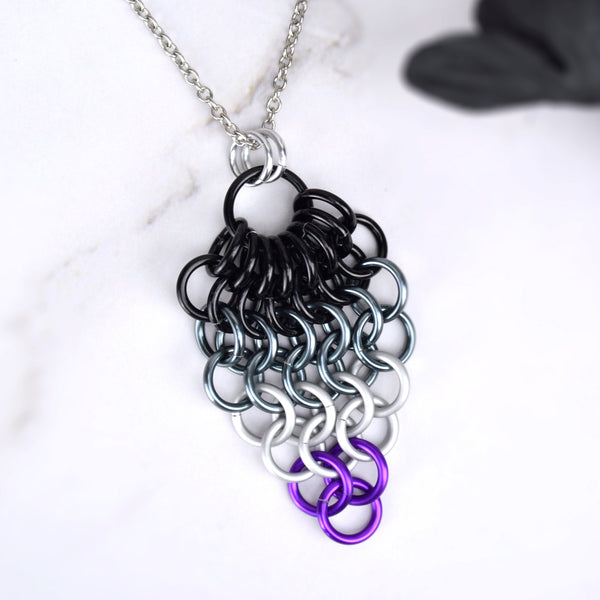 Closeup of short asexual pride chainmaille mesh pendant by Rebeca Mojica displayed on a marbled surface. Pendant is reverse teardrop shape with a large black ring at top. From that ring, a row of 8 smaller black rings hang and are connected to a row of 7 black rings beneath them. Each row gets smaller and smaller: 6 grey rings, 5 grey rings, 4 white rings, 3 white rings, 2 purple rings and 1 purple ring. 