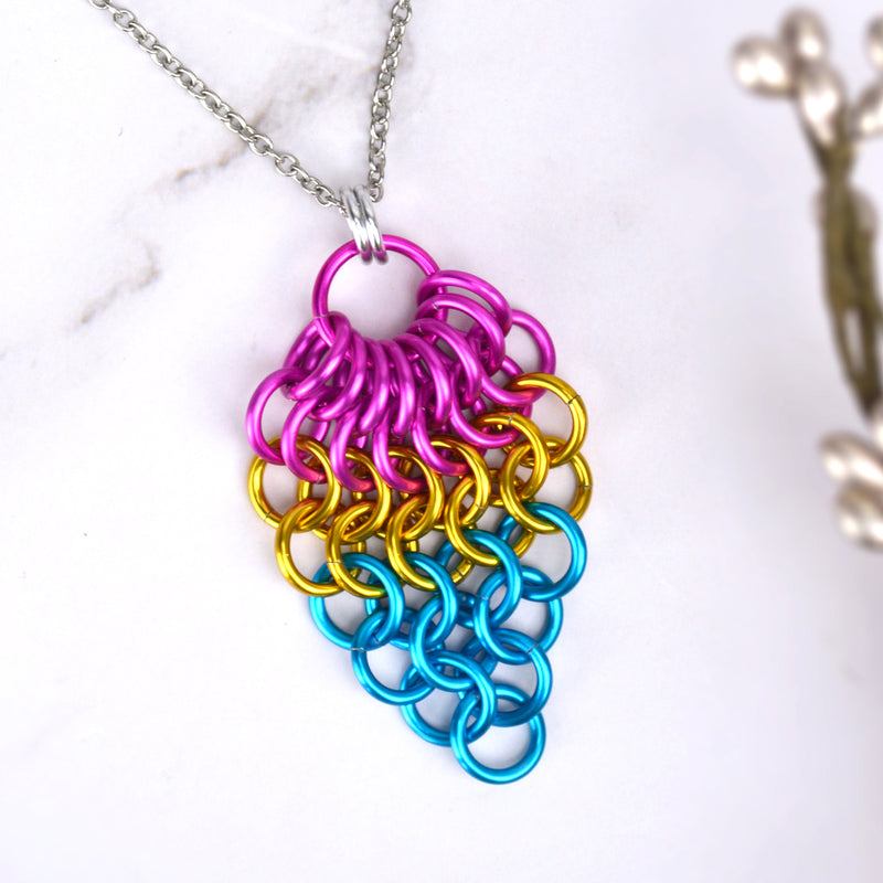 Closeup of short pansexual pride chainmaille mesh pendant by Rebeca Mojica displayed on a marbled surface. Pendant is reverse teardrop shape with a large pink ring at top. From that ring, a row of 8 smaller pink rings hang and are connected to a row of 7 pink rings beneath them. Each row gets smaller and smaller: 6 gold rings, 5 gold rings, 4 blue rings, 3 blue rings, 2 blue rings and 1 blue ring.