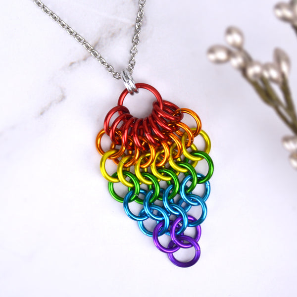 Closeup of short LGBTQ rainbow pride chainmaille mesh pendant by Rebeca Mojica displayed on a marbled surface. Pendant is reverse teardrop shape with a large red ring at top. From that ring, a row of 8 smaller red rings hang and are connected to a row of 7 orange rings beneath them. Each row gets smaller and smaller: 6 yellow rings, 5 green rings, 4 blue rings, 3 blue rings, 2 purple rings and 1 purple ring
