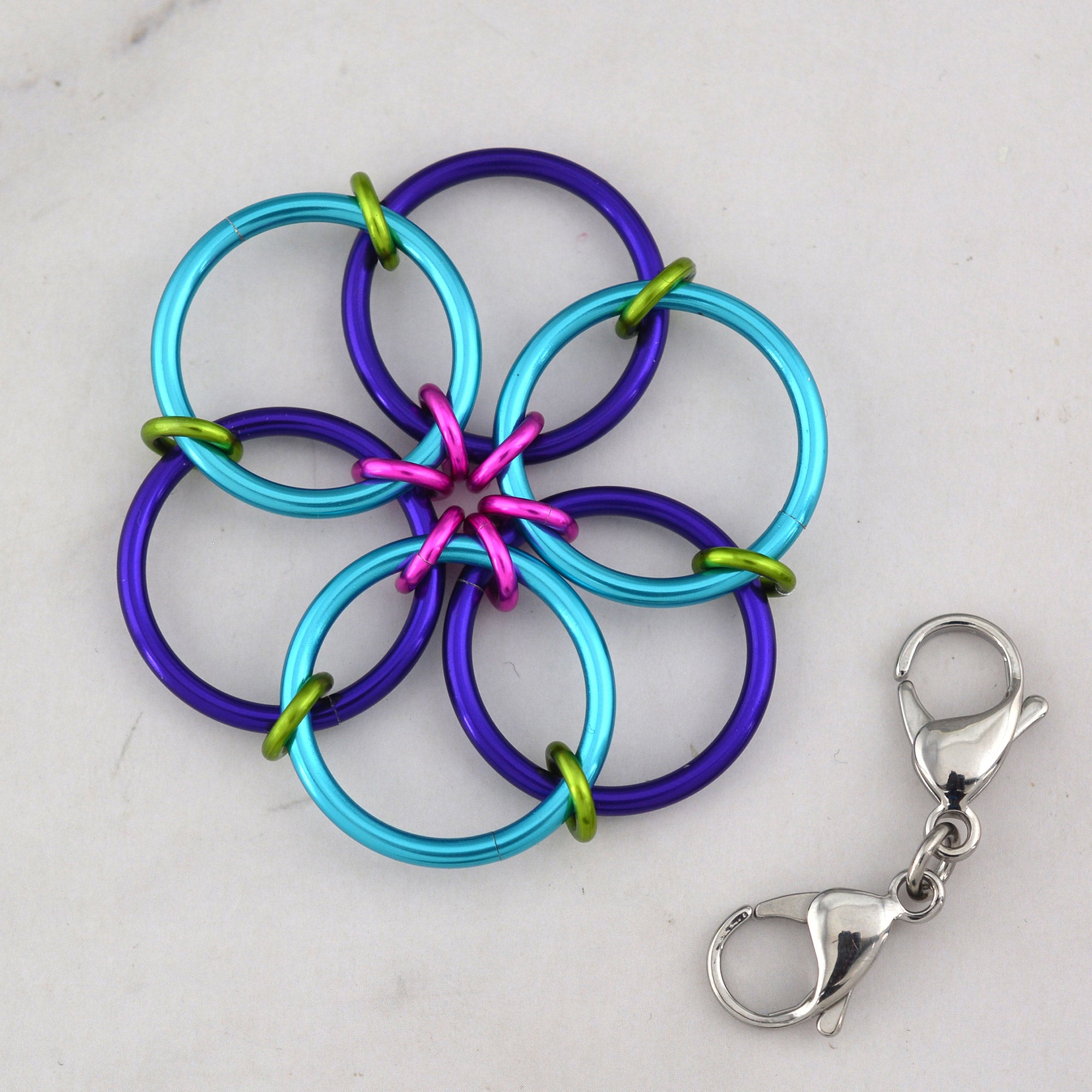 The Funfetti Fidget Flower accessory, made of turquoise, purple, lime green, and hot pink rings, lying flat next to a detached double-ended silver lobster claw clip.