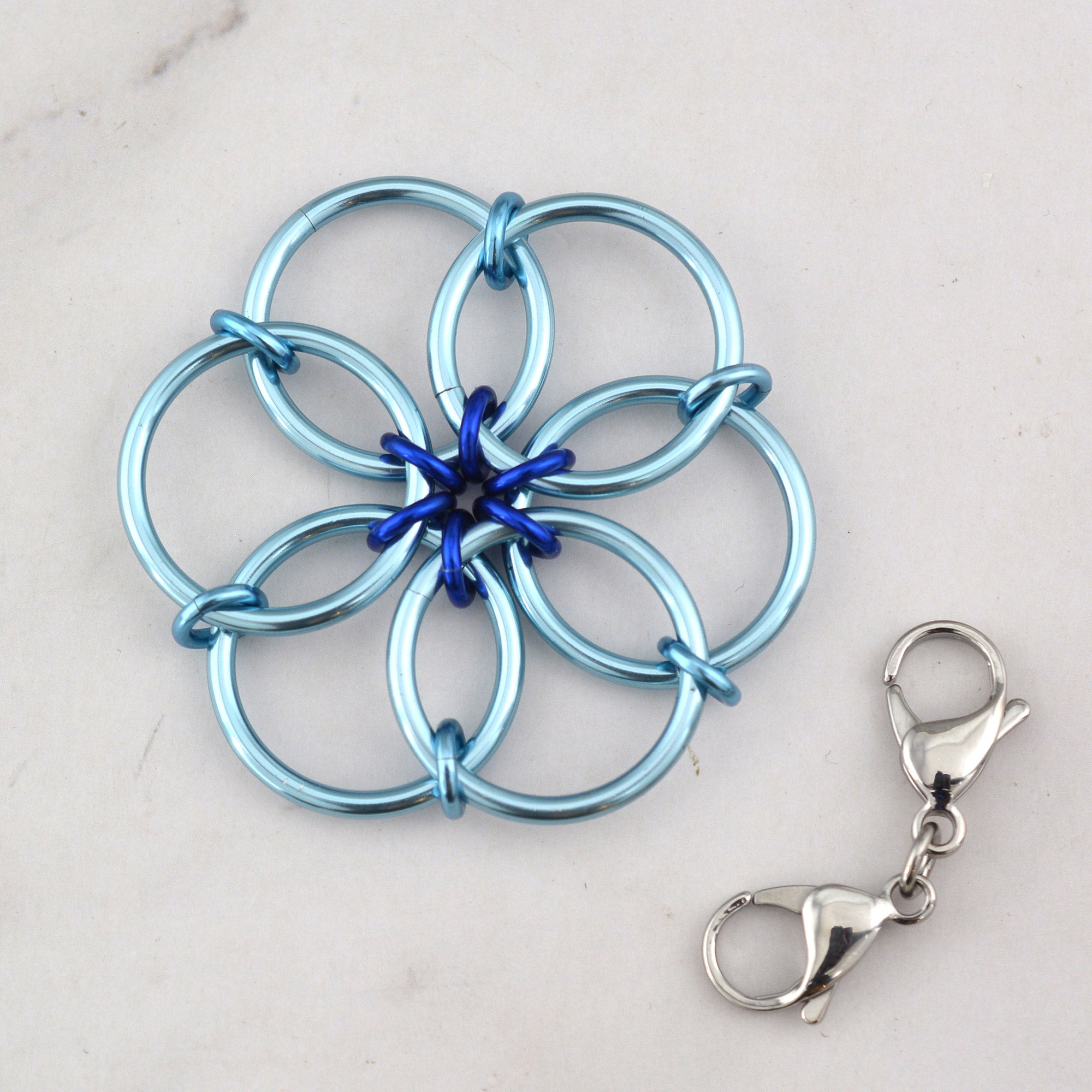 The Light Blue Fidget Flower accessory, made primarily of light blue rings with a royal blue center accent, lying flat next to a detached double-ended stainless steel lobster claw clip.