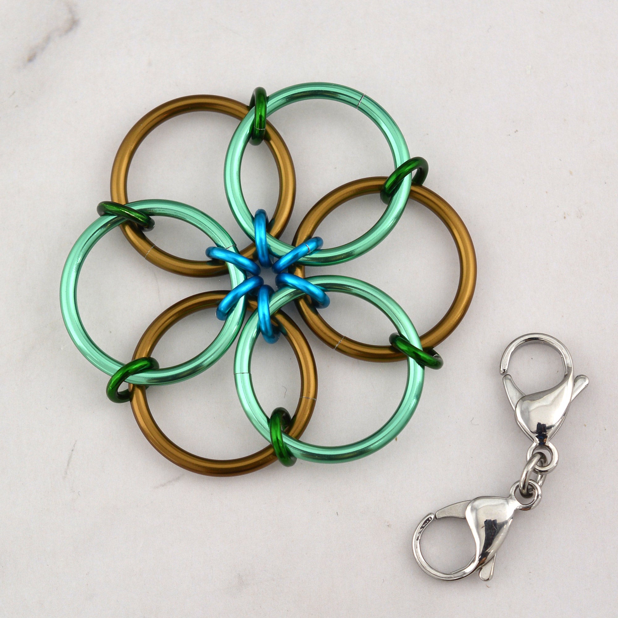 The silver Fidget Flower accessory lying flat next to a detached double-ended steel lobster claw clip, which is used to attach the flower to bags or zippers.