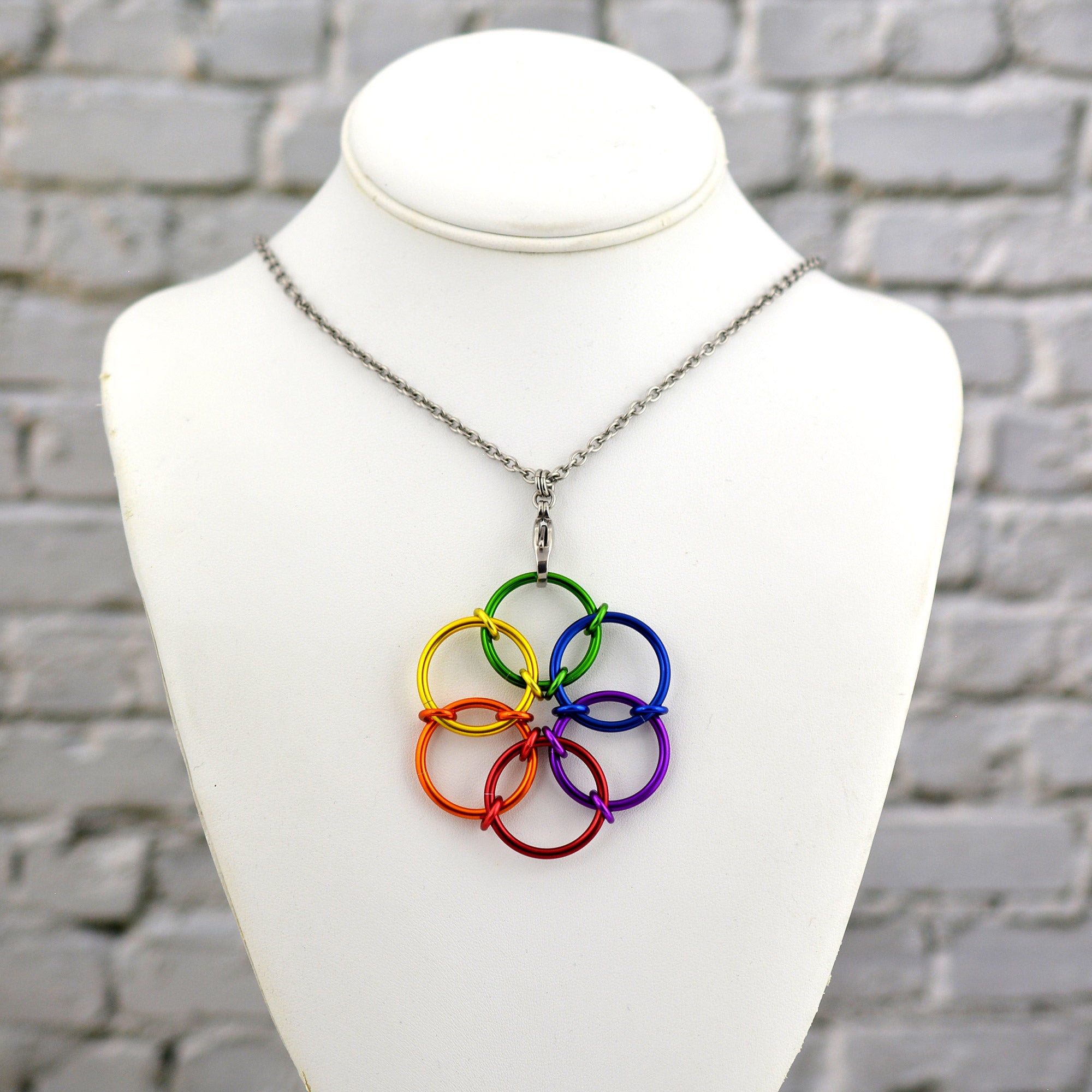 he Rainbow Fidget Flower necklace, featuring the full spectrum of 6 rainbow colors, displayed on a white jewelry neck form against a white brick background.