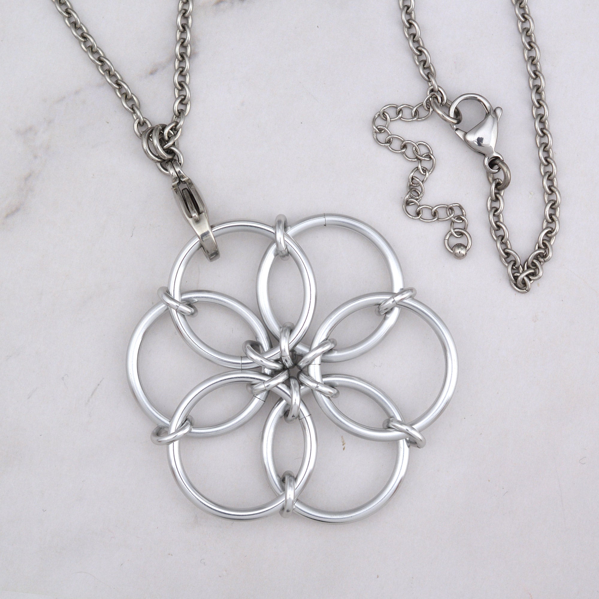 A close-up aerial view of the silver Fidget Flower pendant, showing the detail of the interlocking ring pattern and the chain attached at the top.