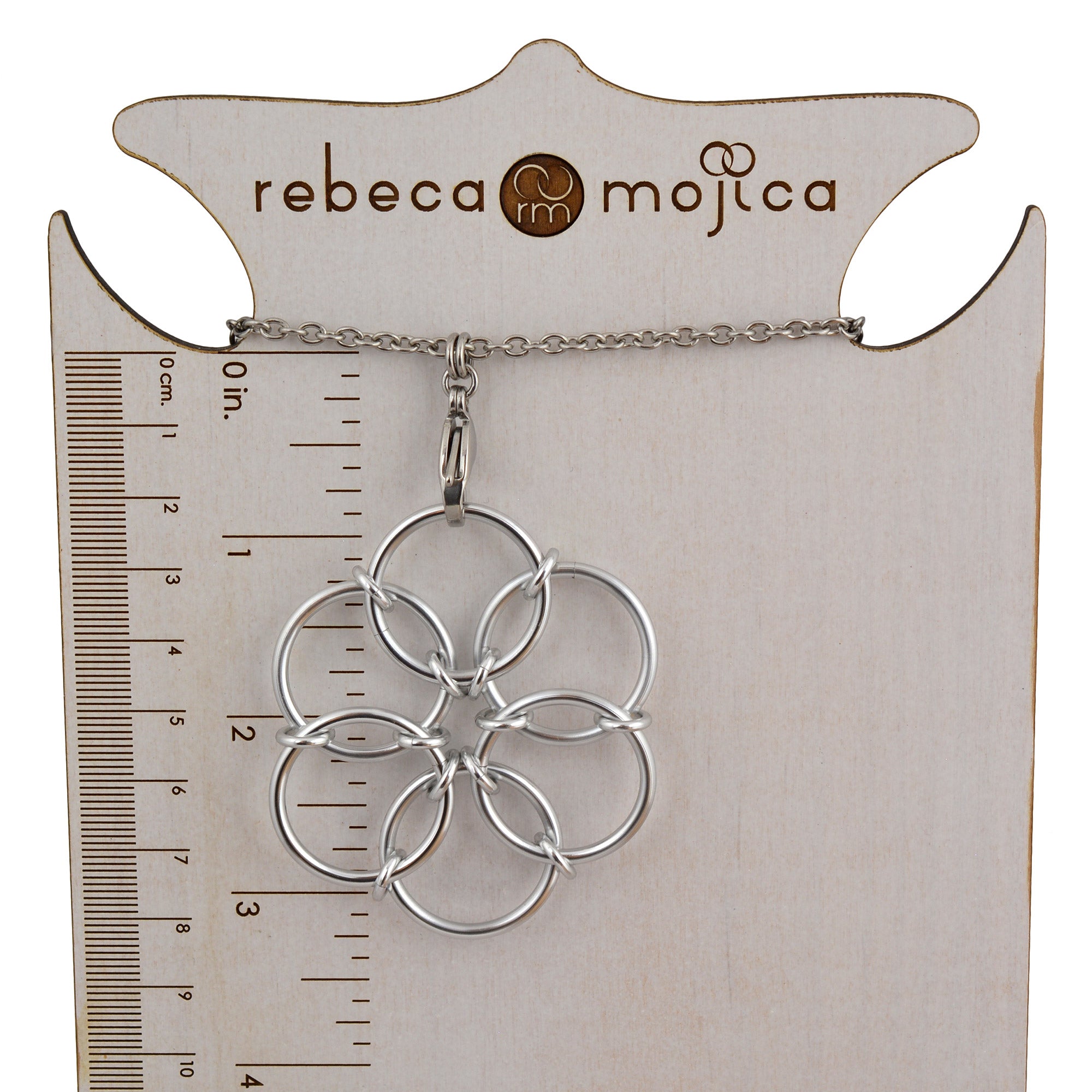 A silver Fidget Flower pendant resting on a branded jewelry display card with a ruler printed on the left side, showing the pendant hangs approximately 3.25" inches from the top of the chain.