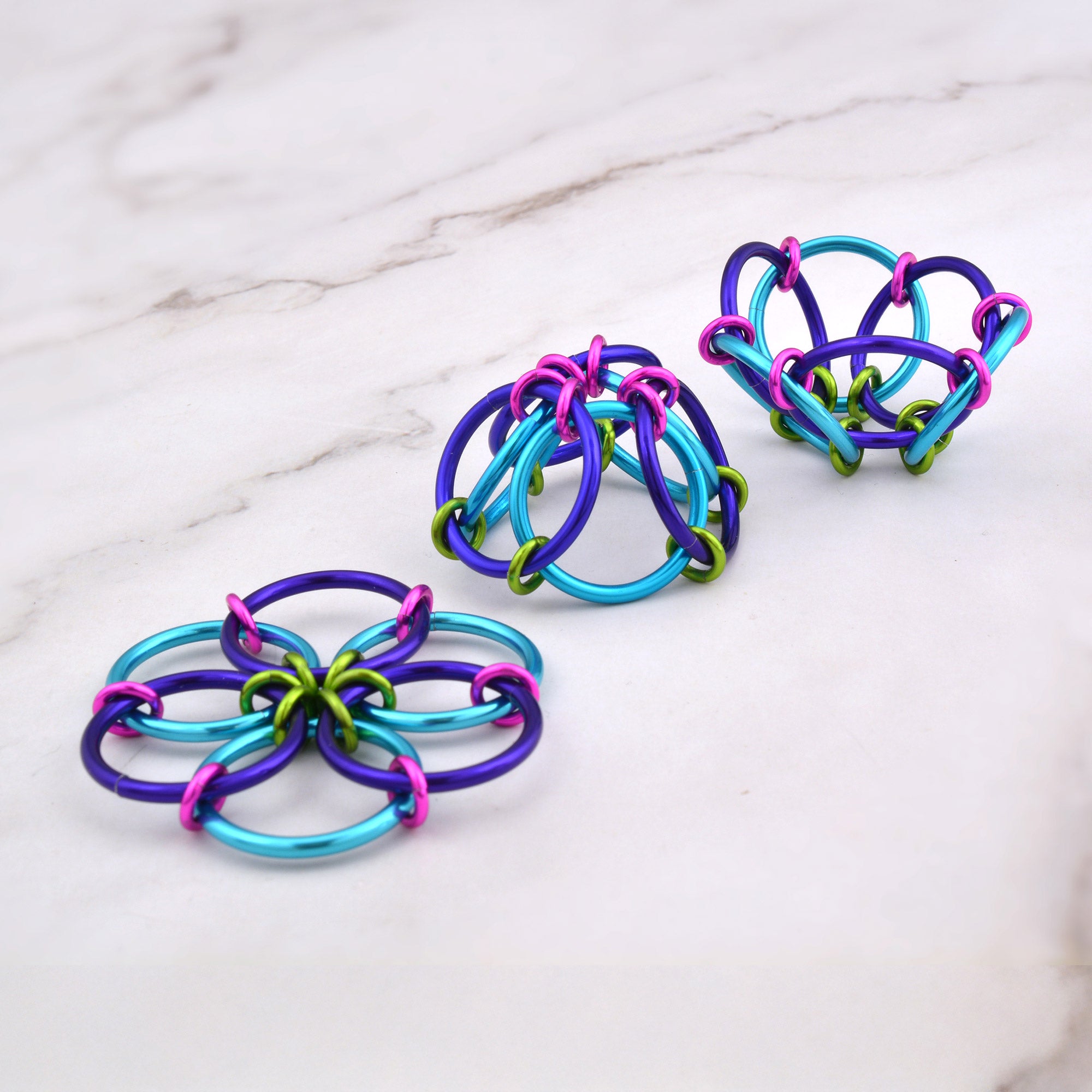A set of three Fidget Flowers in bright turquoise, purple, lime and pink. One is flat in the flower configuration, and the other two are partially folded into 3D dome shapes on a white marble surface.