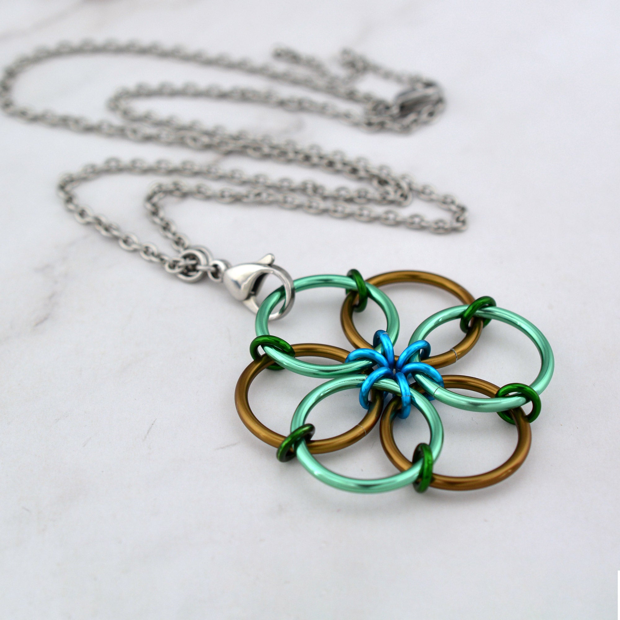 An angled aerial view of the Magical Woodland Fidget Flower necklace lying on a white marble surface, featuring alternating large rings of sea green and russet brown, connected by small forest green and turquoise accent rings.