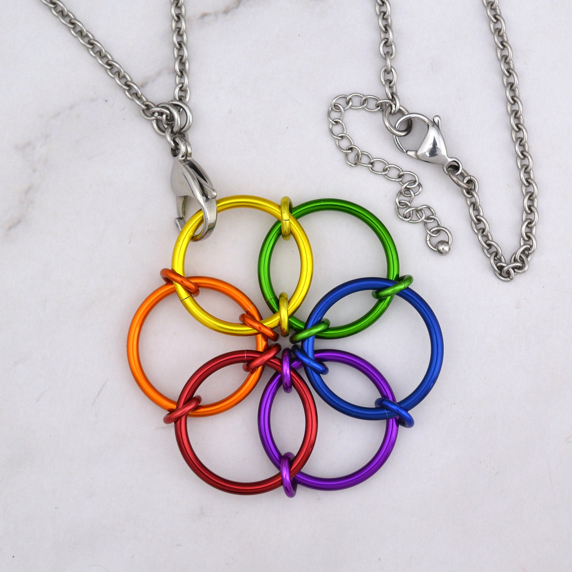 A close up aerial view of the Rainbow Fidget Flower necklace lying on a white marble surface, showing the vibrant rings in the sequence of red, orange, yellow, green, blue, and violet. The steel lobster claw clasp and extender is visible at right.