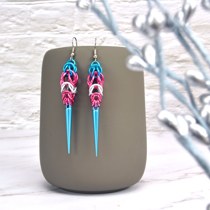 Trans pride chainmaille earrings hanging from a grey tumbler with a white brick background. Earrings are turquoise and pink stripes in the Full Persian weave at the top, followed by white and pink stripes in the Box weave, with a turquoise acrylic spike at the base.