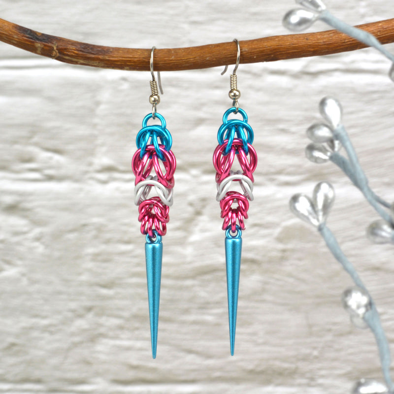 Trans pride chainmaille earrings hanging from a branch. Earrings are turquoise and pink stripes in the Full Persian weave at the top, followed by white and pink stripes in the Box weave, with a turquoise acrylic spike at the base