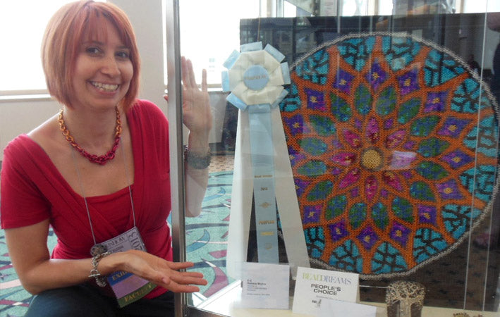 Award-winning chainmaille artist Rebeca Mojica smiling next to her colorful, intricate Chainmaille Mandala piece displayed in a case. The piece is a bead-woven ring design with a white and blue Bead Dreams People's Choice Award ribbon displayed prominently. Rebeca is wearing a red top with a flame ombre knotted metal necklace.
