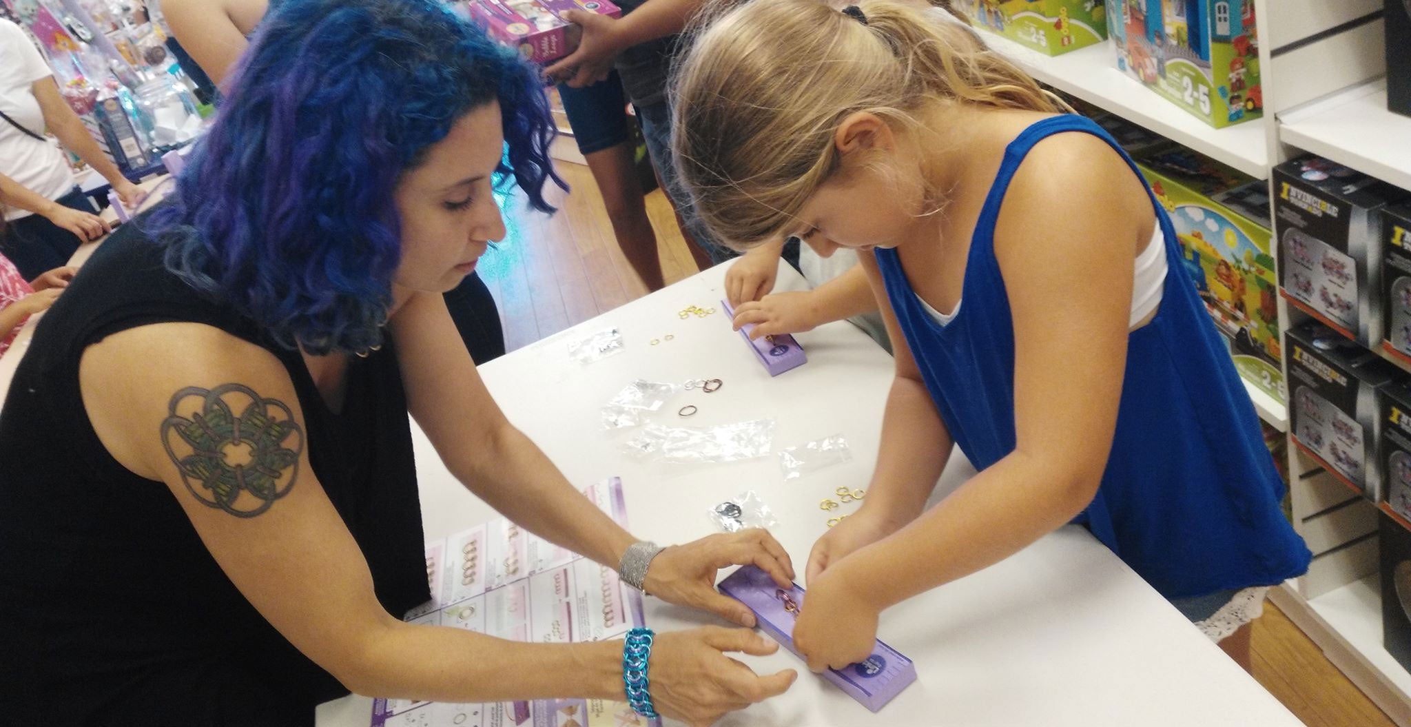 Rebeca Mojica, with blue hair and a chainmaille tattoo, providing in-person chainmaille jewelry instruction to a young student during a creative workshop in a toy shop.