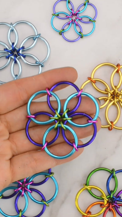 closeup of Rebeca Mojica's hand fidgeting with her brightly colored chainmaille fidget flower. She her thumb and index finger to fold the flower in on itself over and over again. She is wearing sparkly dark blue nail polish and other fidget flowers are visible in the background.
