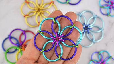 closeup of Rebeca Mojica's hand fidgeting with her brightly colored chainmaille fidget flower. She her thumb and index finger to fold the flower in on itself over and over again. She is wearing sparkly dark blue nail polish and other fidget flowers are visible in the background.