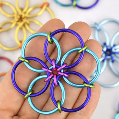 closeup of Rebeca Mojica's hand fidgeting with her brightly colored chainmaille fidget flower. She her thumb and index finger to fold the flower in on itself over and over again. She is wearing sparkly dark blue nail polish and other fidget flowers are visible in the background.